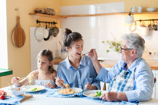 Happy multigenerational family eating pasta together at home kitchen, having fun. Grandmother feeding adult daughter, child enjoying comfort food. Candid, authentic lifestyle, joyful mealtime moment.
